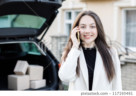 Young business woman standing at the car with parcels, coming home by car. The girl is standing and talking on mobile phone near the car. Concept of buying goods online and delivering them home 117193491