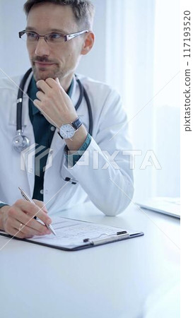 Doctor and a patient. The physician, wearing a white medical coat over a green shirt, gesturing with his hands during a consultation in the clinic. Medicine concept Doctor and a patient. The physician, wearing a white medical coat over a green shirt, gesturing with his hands during a consultation in the clinic. Medicine concept 117193520