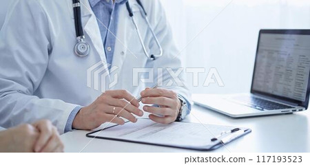 A doctor and a patient. The physician, wearing a white medical coat over a blue shirt, is filling out a medical record form during a consultation in the clinic. Medical service A doctor and a patient. The physician, wearing a white medical coat over a blue shirt, is filling out a medical record form during a consultation in the clinic. Medical service 117193523