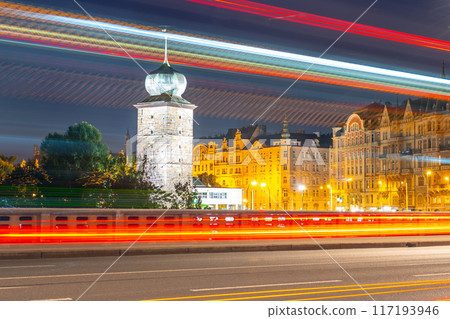 A long exposure photograph captures the vibrant lights of Prague at dusk, showcasing the Sitkov Water Tower and the Masaryk Embankment. Traffic streaks illuminate the road in front of the buildings. 117193946