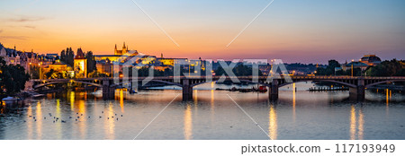 Jirasek bridge crosses the Vltava River in Prague, with Prague Castle illuminated on the opposite bank. The setting sun casts a warm glow on the cityscape, creating a serene and picturesque scene. Jirasek bridge crosses the Vltava River in Prague, with Prague Castle illuminated on the opposite bank. The setting sun casts a warm glow on the cityscape, creating a serene and picturesque scene. 117193949