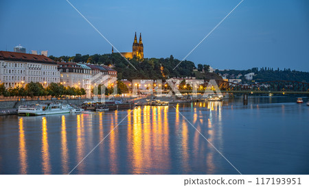 A view of Prague iconic Vysehrad Castle, illuminated against the twilight sky, as it sits atop a hill overlooking the Vltava River. The Rasin Embankment stretches along the riverbank, dotted with 117193951