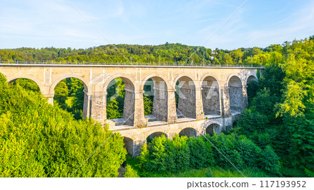 The Sychrov Railway Bridge stands majestically with graceful arches, surrounded by lush greenery. The bright sky enhances the tranquil setting. 117193952