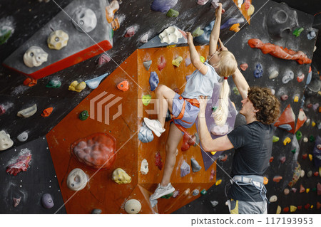 Male instructor is helping a little girl in climbing gym to climb on artificial rocks on wall. Little girl is engaged in rock climbing in the children's entertainment center. Blonde caucasian girl Male instructor is helping a little girl in climbing gym to climb on artificial rocks on wall. Little girl is engaged in rock climbing in the children's entertainment center. Blonde caucasian girl 117193953