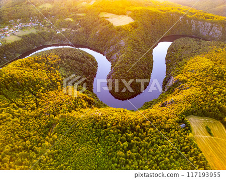 The Vltava River weaves gracefully through lush green landscapes in Czechia, highlighted by vibrant hues of summer. This serene waterway reflects the golden glow of the setting sun, offering a 117193955