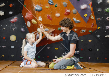 Male instructor is talking to little girl in climbing gym explaining safety requirements showing artificial rocks on wall. Little girl is engaged in rock climbing in the children's entertainment Male instructor is talking to little girl in climbing gym explaining safety requirements showing artificial rocks on wall. Little girl is engaged in rock climbing in the children's entertainment 117193956