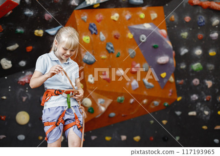 Little girl standing in rock-climbing gym. Girl is ready to climb up artificial wall wearing sports outfit and safety equipment. Blonde girl wearing shorts and t-shirt. 117193965
