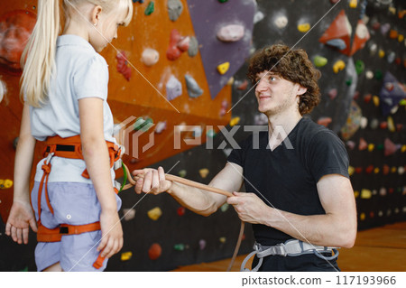 Instructor man is helping client small girl to put on safety harnesses in rock climbing center indoors. Little girl is engaged in rock climbing in the children's entertainment center. Blonde caucasian Instructor man is helping client small girl to put on safety harnesses in rock climbing center indoors. Little girl is engaged in rock climbing in the children's entertainment center. Blonde caucasian 117193966