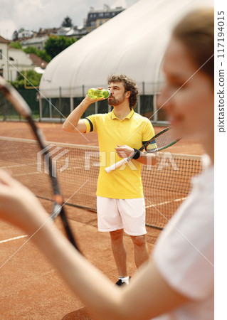 Photo of male tennis player and female in front of him. Man standing on a court and drinking a water. Man wearing yellow t-shirt and white shorts. 117194015