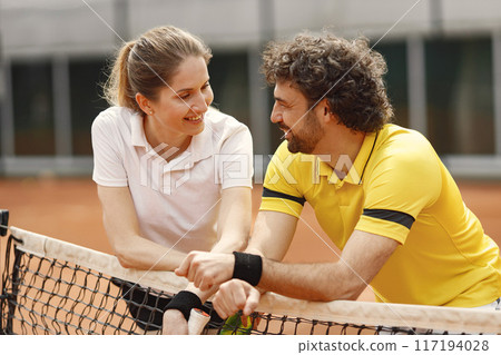 Curly man and woman tennis players standing on open summer court with rackets in hands. Couple friendly chatting after the match while leaning on a net. Man wearing yellow t-shirt and woman white one. Curly man and woman tennis players standing on open summer court with rackets in hands. Couple friendly chatting after the match while leaning on a net. Man wearing yellow t-shirt and woman white one. 117194028