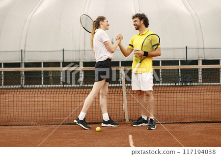 Curly man and woman tennis players standing on open summer court with rackets and balls in hands. Couple friendly shaking hands at court before the match. Man wearing yellow t-shirt and woman the Curly man and woman tennis players standing on open summer court with rackets and balls in hands. Couple friendly shaking hands at court before the match. Man wearing yellow t-shirt and woman the 117194038