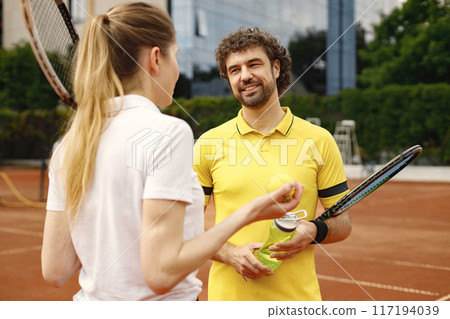 Curly man and woman tennis players standing on open summer court with rackets and balls in hands. Couple friendly chatting before the match. Man wearing yellow t-shirt and holding a bottle with water. Curly man and woman tennis players standing on open summer court with rackets and balls in hands. Couple friendly chatting before the match. Man wearing yellow t-shirt and holding a bottle with water. 117194039