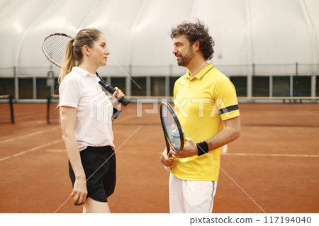 Curly man and woman tennis players standing on open summer court with rackets and balls in hands. Couple friendly chatting bedore the match. Man wearing yellow t-shirt and woman white one. 117194040