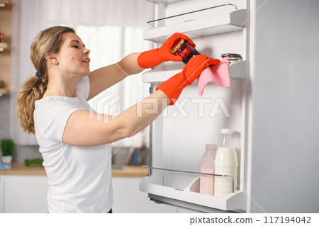 Portrait of a woman cleaning in a kitchen and wearing red gloves. Caucasian woman doing housework. Adult woman washing a refrigerator. Portrait of a woman cleaning in a kitchen and wearing red gloves. Caucasian woman doing housework. Adult woman washing a refrigerator. 117194042