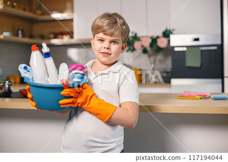 Portrait of a blonde boy cleaning in a kitchen and wearing orange gloves. Caucasian boy helping his mother to do housework. Boy holding blue busket with different rugs and detergents. Portrait of a blonde boy cleaning in a kitchen and wearing orange gloves. Caucasian boy helping his mother to do housework. Boy holding blue busket with different rugs and detergents. 117194044