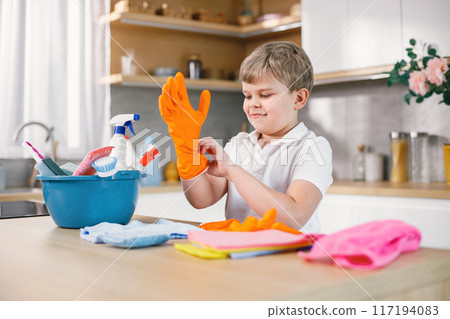 Blonde boy cleaning in a kitchen and wearing orange gloves. Caucasian boy helping his mother to do housework. Blue busket with different rugs and detergents on a table. 117194083