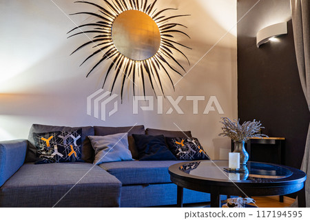 Tiny living room with sofa, coffee table and wall decor in form of sun. Cozy interior in warm Tiny living room with sofa, coffee table and wall decor in form of sun. Cozy interior in warm 117194595