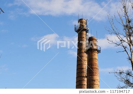 Two tall brick chimneys above abandoned boiler building for venting smoke, combustion byproducts from factory heating. Industrial red towers or pipe with mobile antennas. Smokestacks with cell site Two tall brick chimneys above abandoned boiler building for venting smoke, combustion byproducts from factory heating. Industrial red towers or pipe with mobile antennas. Smokestacks with cell site 117194717