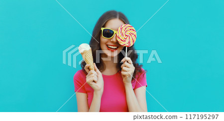 Summer portrait happy young woman holding ice cream cone, lollipop blowing kiss on blue background Summer portrait happy young woman holding ice cream cone, lollipop blowing kiss on blue background 117195297