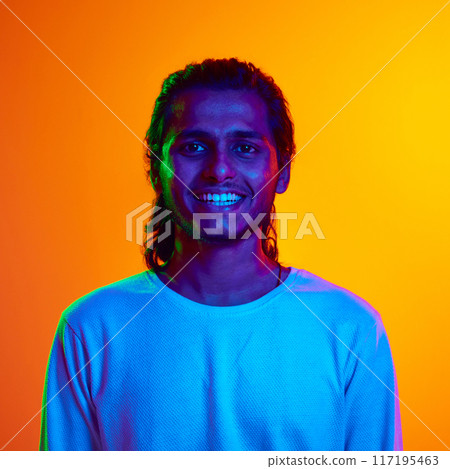 Portrait of young Indian man dressed casually looks at camera with light smile in neon light against gradient orange background. 117195463