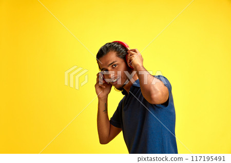Young middle eastern man wearing headphones looking at camera to listen music against yellow studio background. 117195491