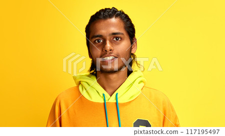 Portrait of young Indian man dressed stylish, vibrant hoodie and looking at camera positive smiling against yellow studio background. Portrait of young Indian man dressed stylish, vibrant hoodie and looking at camera positive smiling against yellow studio background. 117195497