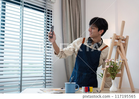 Young Artist Taking a Selfie in Studio with Easel and Art Supplies, Wearing Apron and Headphones 117195565