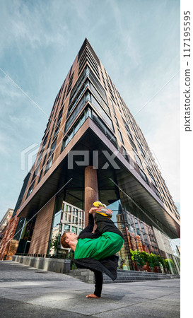 Urban breakdancer in stylish outfit flips upside down in thrilling handstand in city with buildings around him. 117195595