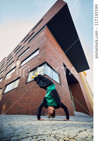 Dancer defies gravity with upside-down handstand flip, vibrant yellow shoes pointing skyward, framed by towering urban architecture. Dancer defies gravity with upside-down handstand flip, vibrant yellow shoes pointing skyward, framed by towering urban architecture. 117195599