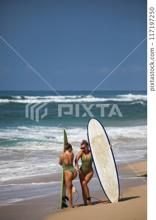 Two beautiful blondes in swimsuits pose with surfboards on the beach 117197250