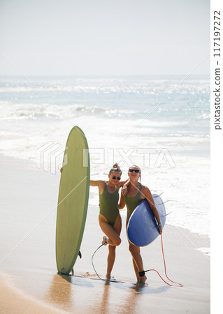 Two beautiful blondes in swimsuits pose with surfboards on the beach Two beautiful blondes in swimsuits pose with surfboards on the beach 117197272