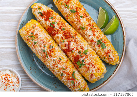 Elotes mexican street corn cob covered in spices and cheese being served on plate Elotes mexican street corn cob covered in spices and cheese being served on plate 117197805