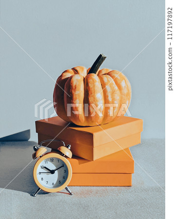 Large pumpkin in square podium with orange alarm clock. Still life in monochrome palette. Pumpkin time concept. Halloween and Thanksgiving decoration Large pumpkin in square podium with orange alarm clock. Still life in monochrome palette. Pumpkin time concept. Halloween and Thanksgiving decoration 117197892