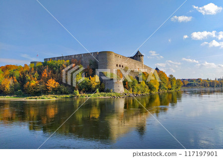 Scenic view of Ivangorod Fortress surrounded by colorful autumn trees, reflecting in calm river below under clear blue sky 117197901