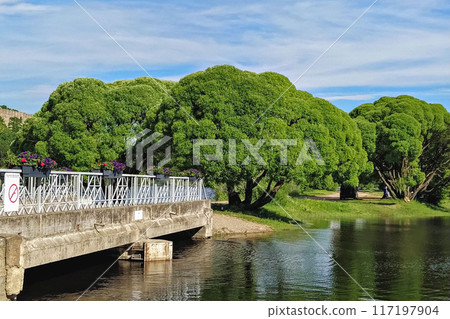 Charming bridge decorated with flowers spans over river, with lush green trees in background under partly cloudy sky 117197904