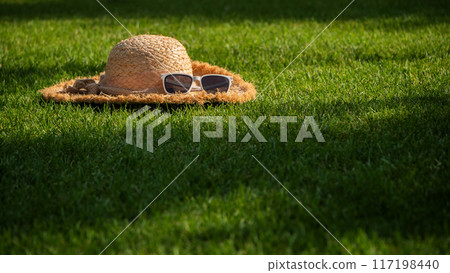 A straw hat adorned with white sunglasses sits on vibrant green grass, with purple flowers blooming in the background, symbolizing a peaceful summer afternoon. 117198440