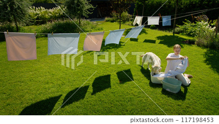 A girl hangs laundry on a clothesline in a sunlit backyard, accompanied by a Golden Retriever dog, creating a peaceful and homey scene. 117198453