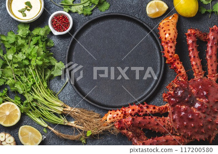 Top view of fresh king crab legs, herbs, and lemon wedges surrounding an empty black plate on a dark textured background, ready for a gourmet seafood preparation Top view of fresh king crab legs, herbs, and lemon wedges surrounding an empty black plate on a dark textured background, ready for a gourmet seafood preparation 117200580