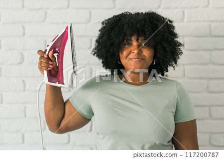 Young satisfied african american housekeeper woman wear mint t-shirt hold in hands on iron on brick wall background. Housework housekeeping concept Young satisfied african american housekeeper woman wear mint t-shirt hold in hands on iron on brick wall background. Housework housekeeping concept 117200787