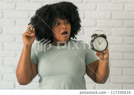 Surprised african american woman holding alarm clock amazed with open mouth and surprise happy face at home, on brick background. Being late and time passing quickly concept 117200816