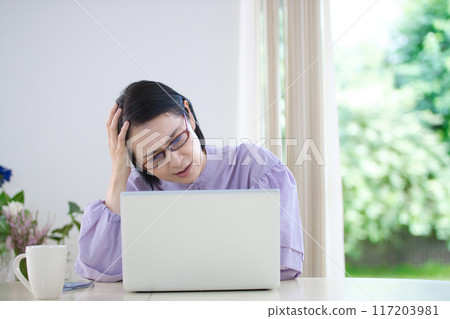 A middle-aged woman holding her head while looking at a computer in the living room 117203981