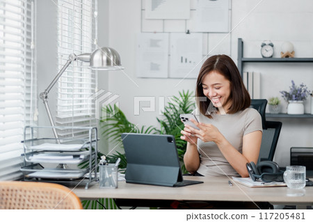 A young woman in a home office setting, smiling while using her smartphone. The workspace includes a laptop, documents, and modern decor. 117205481