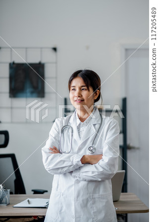 A confident female doctor in a white coat with a stethoscope stands in a modern medical office, ready to assist patients. A confident female doctor in a white coat with a stethoscope stands in a modern medical office, ready to assist patients. 117205489