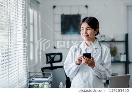 A young female doctor in a white coat using a smartphone in a modern medical office with bright natural light coming through the window. A young female doctor in a white coat using a smartphone in a modern medical office with bright natural light coming through the window. 117205490
