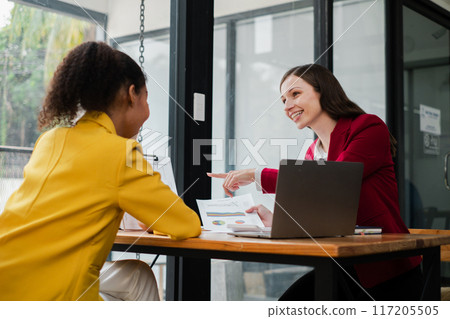 Two women engaged in a business meeting, discussing documents in a modern office environment with laptops on the table. 117205505