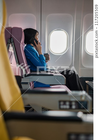Businesswoman in blue blazer talking on phone and holding coffee cup while seated on an airplane during flight. 117205509