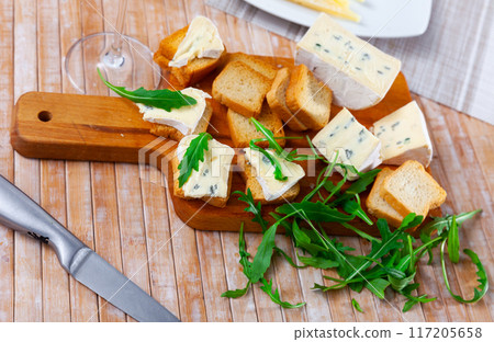On table there is cutting board with blue cheese and hardtack decorated with arugula On table there is cutting board with blue cheese and hardtack decorated with arugula 117205658