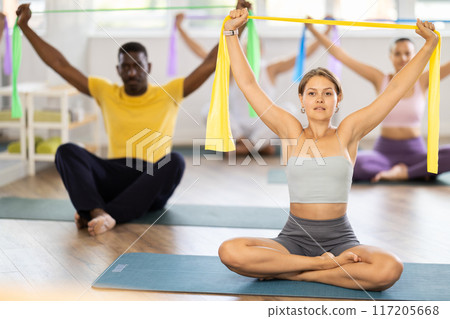 Girl conducts Pilates multinational group sitting with elastic band in hands above head body bends Girl conducts Pilates multinational group sitting with elastic band in hands above head body bends 117205668