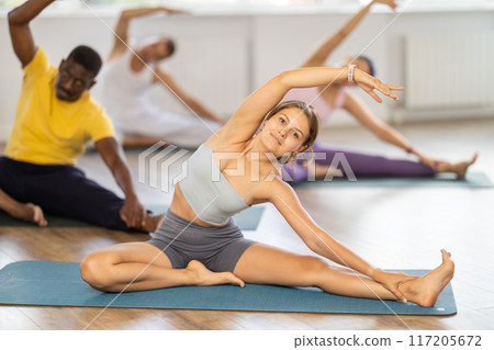 Young female teacher working out Parivritta Janu Shirshasana with multinational students group Young female teacher working out Parivritta Janu Shirshasana with multinational students group 117205672