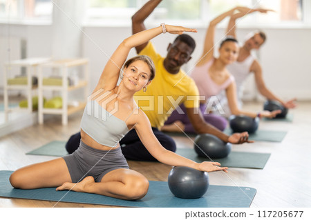 Multinational group of people lie on mats, hold Pilates ball between their knees and perform abdominal exercises during group exercise in fitness studio 117205677